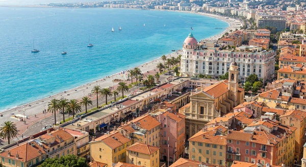 Photographie réaliste de la ville de Nice, France, montrant des éléments architecturaux et urbains caractéristiques : vue sur la Promenade des Anglais avec ses bâtiments emblématiques, ou une perspective des quartiers reconnaissables de Nice (vieille ville avec ses façades colorées, front de mer, ou environnement urbain côtier typique). La photo doit être prise de jour, bien éclairée, avec une qualité professionnelle, sans texte ni logo visible. Elle doit représenter distinctement Nice (non simplement une ville côtière générique) pour se démarquer visuellement des images existantes pour Cannes et Antibes.
